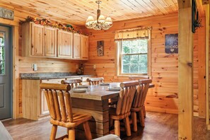 A wooden cabin interior with a dining table, chairs, a chandelier, and a window with curtains.