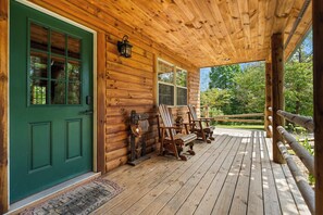 Eine Holzhütte mit grüner Tür, einer Veranda mit Holzstühlen und Blick auf den umliegenden Wald.