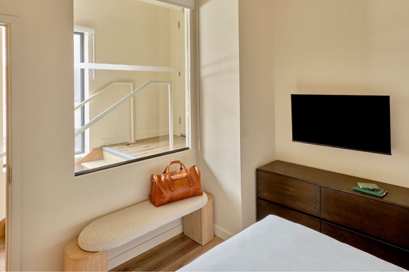 Quarto de hotel com um espelho grande, um banco, uma TV e uma cômoda de madeira.