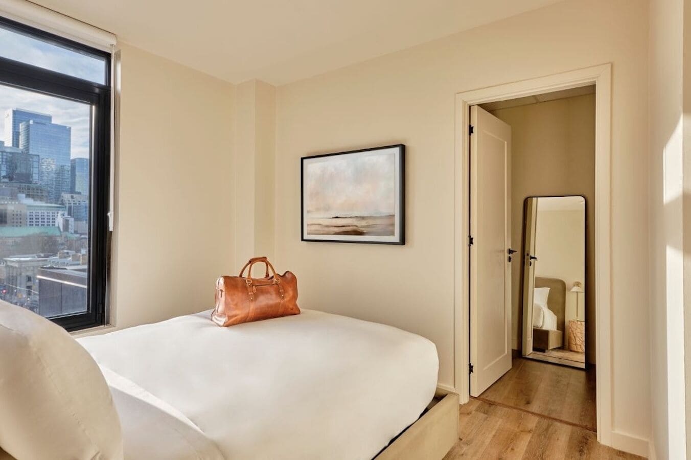 Una habitació d'hotel amb un llit, una finestra amb vista a la ciutat, un quadre al mur i una porta que condueix a un bany.