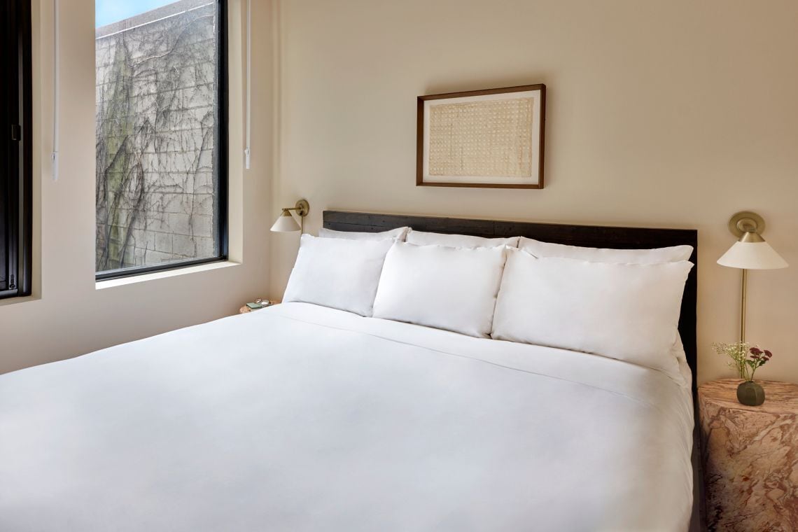 A neatly made bed with white linens, a framed artwork above it, and a lamp on a side table.
