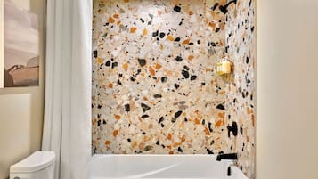 Un baño con piso de travertino, una bañera blanca y un aplique de pared.
