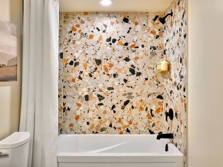 Un baño con piso de travertino, una bañera blanca y un aplique de pared.