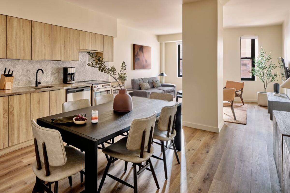 Eine moderne Küche mit Holzschränken, ein Essbereich mit dunklem Tisch und hellen Stühlen sowie ein Wohnbereich mit grauem Sofa und Couchtisch.