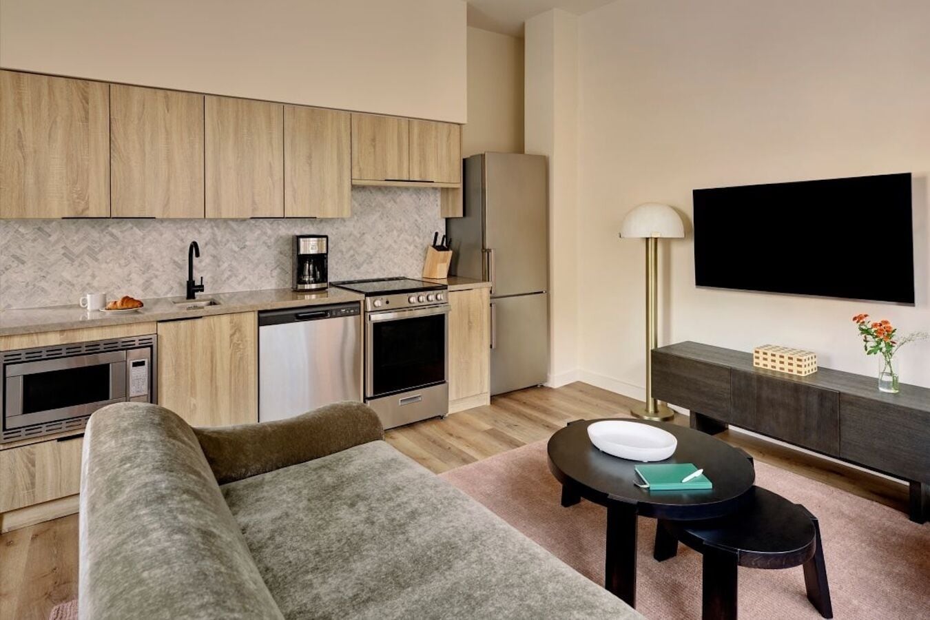 A modern kitchen with wooden cabinets, a built-in microwave, and a coffee maker. There is a dining area with a black table and a sofa. A flat-screen TV is mounted on the wall above the sofa.