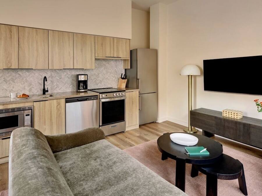 A modern kitchen with wooden cabinets, a built-in microwave, and a coffee maker. There is a dining area with a black table and a sofa. A flat-screen TV is mounted on the wall above the sofa.
