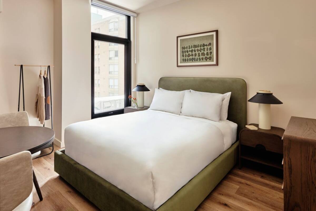 Ein ordentlich eingerichtetes Hotelzimmer mit einem großen Bett, einem kleinen runden Tisch, einem Sessel und einem Fenster mit Blick auf die Stadt.