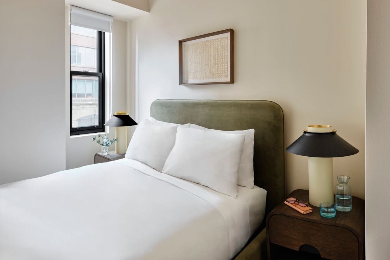 Una cama bien tendida con sábanas blancas, un cabecero tapizado en verde y dos mesitas de noche con lámparas y vasos.