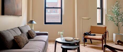 Una sala de estar con sofá, una mesa redonda, una silla y una ventana con vistas a un edificio.