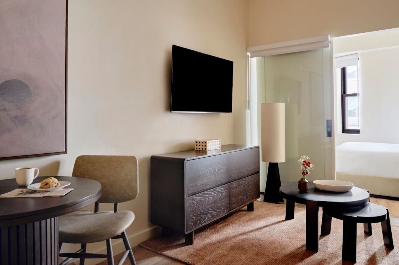 A modern hotel room with a flat-screen TV, a dark wooden dresser, a round dining table with two chairs, a small side table with a vase and a bowl, and a large window with a view of the outdoors.