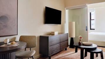 A modern hotel room with a flat-screen TV, a dark wooden dresser, a round dining table with two chairs, a small side table with a vase and a bowl, and a large window with a view of the outdoors.