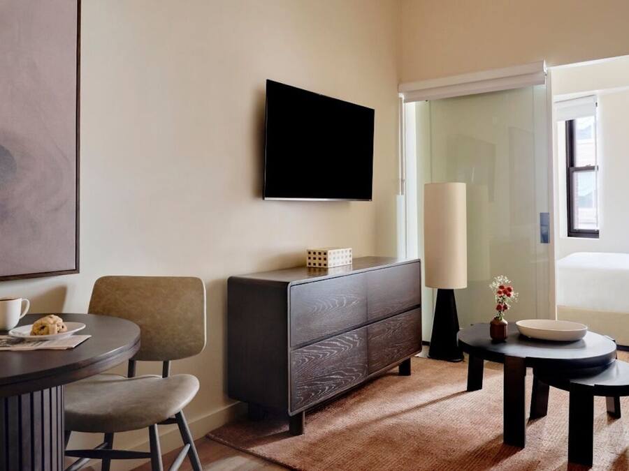 A modern hotel room with a flat-screen TV, a dark wooden dresser, a round dining table with two chairs, a small side table with a vase and a bowl, and a large window with a view of the outdoors.