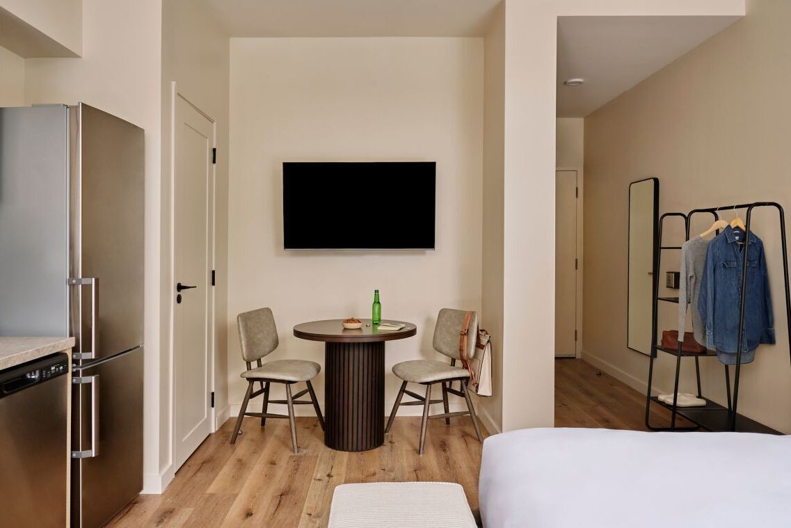 Une chambre d’hôtel comprenant un lit, une table à manger avec deux chaises, un téléviseur fixé au mur, un porte-vêtements et un réfrigérateur.
