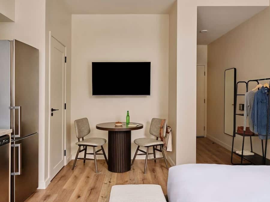 Une chambre d’hôtel comprenant un lit, une table à manger avec deux chaises, un téléviseur fixé au mur, un porte-vêtements et un réfrigérateur.