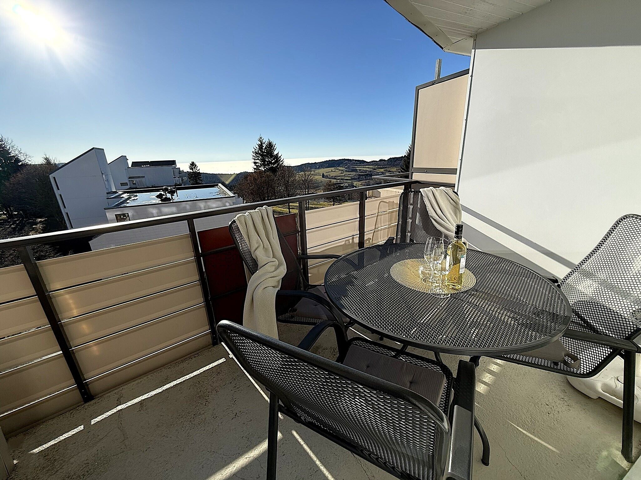 A balcony with a table and chairs, a bottle of wine, and a view of a swimming pool and buildings.