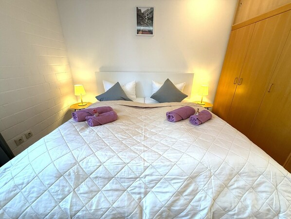 A neatly made bed with a quilted white duvet, two pillows, and two rolled towels on the bed. There are two bedside lamps on either side of the bed.