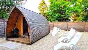 Una cabaña acogedora de madera con techo curvo, un pequeño porche con una silla, y una zona de estar con sillones blancos.