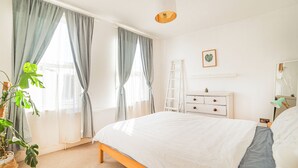 Un dormitorio con cama, cómoda, escalera, planta y ventana con cortinas.