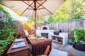 Un patio al aire libre con mesa de madera, sillas, una parrilla y un horno para pizza.