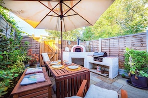 Eine Terrasse im Freien mit einem Holztisch, Stühlen, einem Grill und einem Pizzaofen.