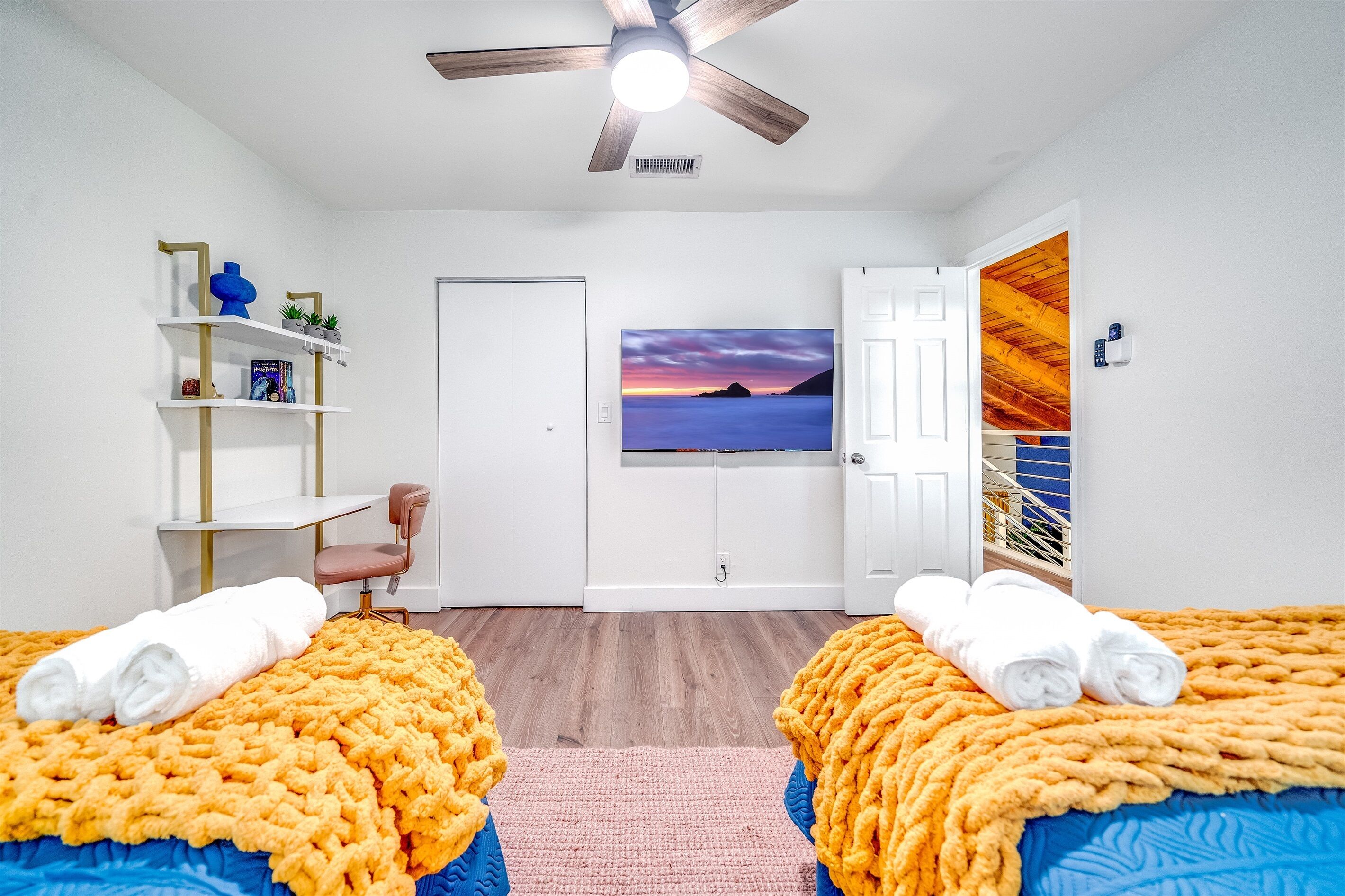 A hotel room with two beds, a desk, a chair, a ceiling fan, a painting, and a view of a wooden structure.