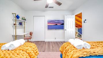 A hotel room with two beds, a desk, a chair, a ceiling fan, a painting, and a view of a wooden structure.