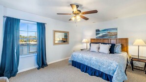 A bedroom with a bed, a ceiling fan, a window with curtains, a painting on the wall, and a nightstand with a lamp. - Blue Bungalow on 1st! (Cedar Key)