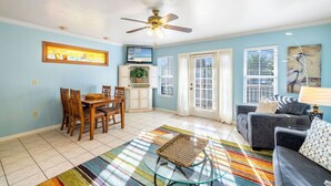 A living room with a dining area, a ceiling fan, a TV, and a window with blinds. - Blue Bungalow on 1st! (Cedar Key)