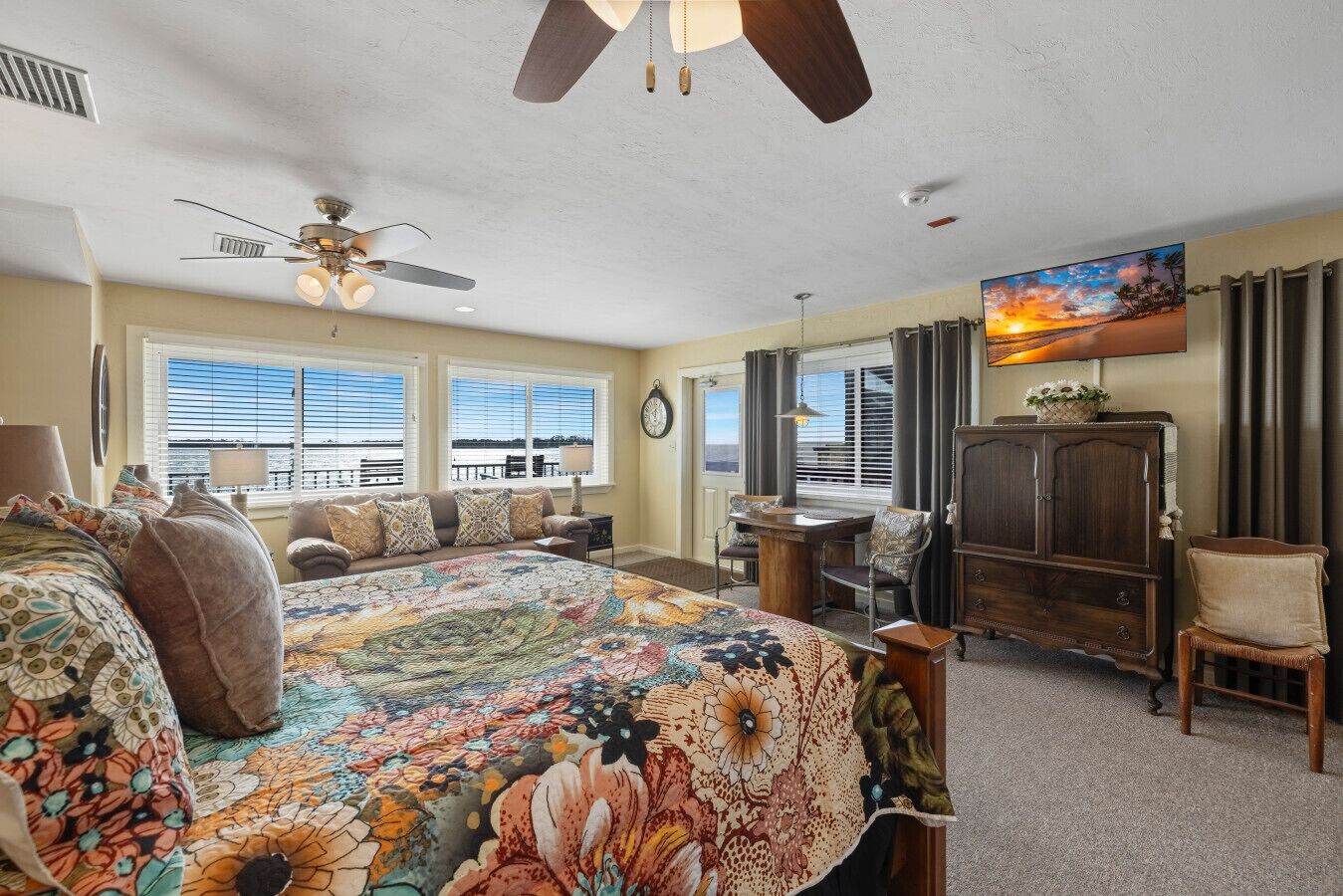 Un dormitorio con una cama grande, un ventilador de techo, un escritorio y vista al océano a través de las ventanas.