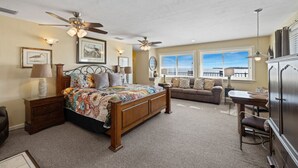 A bedroom with a large bed, a ceiling fan, a sofa, a desk, and a view of the ocean through the windows. - Corrigan`s Reef at Harbour Master Suites (Cedar Key)