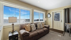 A living room with a brown leather sofa, two lamps, a clock, and a view of the ocean through large windows. - Corrigan`s Reef at Harbour Master Suites (Cedar Key)