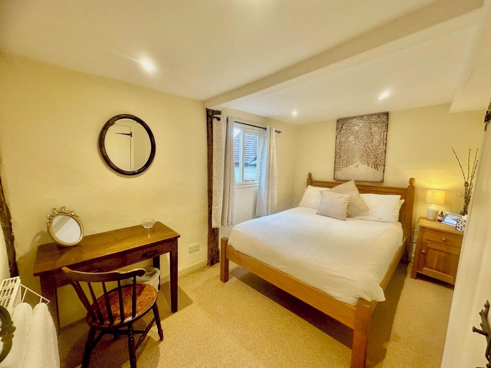 A bedroom with a bed, a desk, a chair, a mirror, a window, and a nightstand with a lamp.