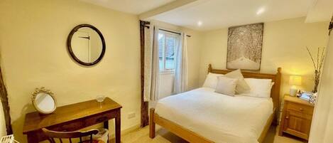 A bedroom with a bed, a desk, a chair, a mirror, a window, and a nightstand with a lamp.