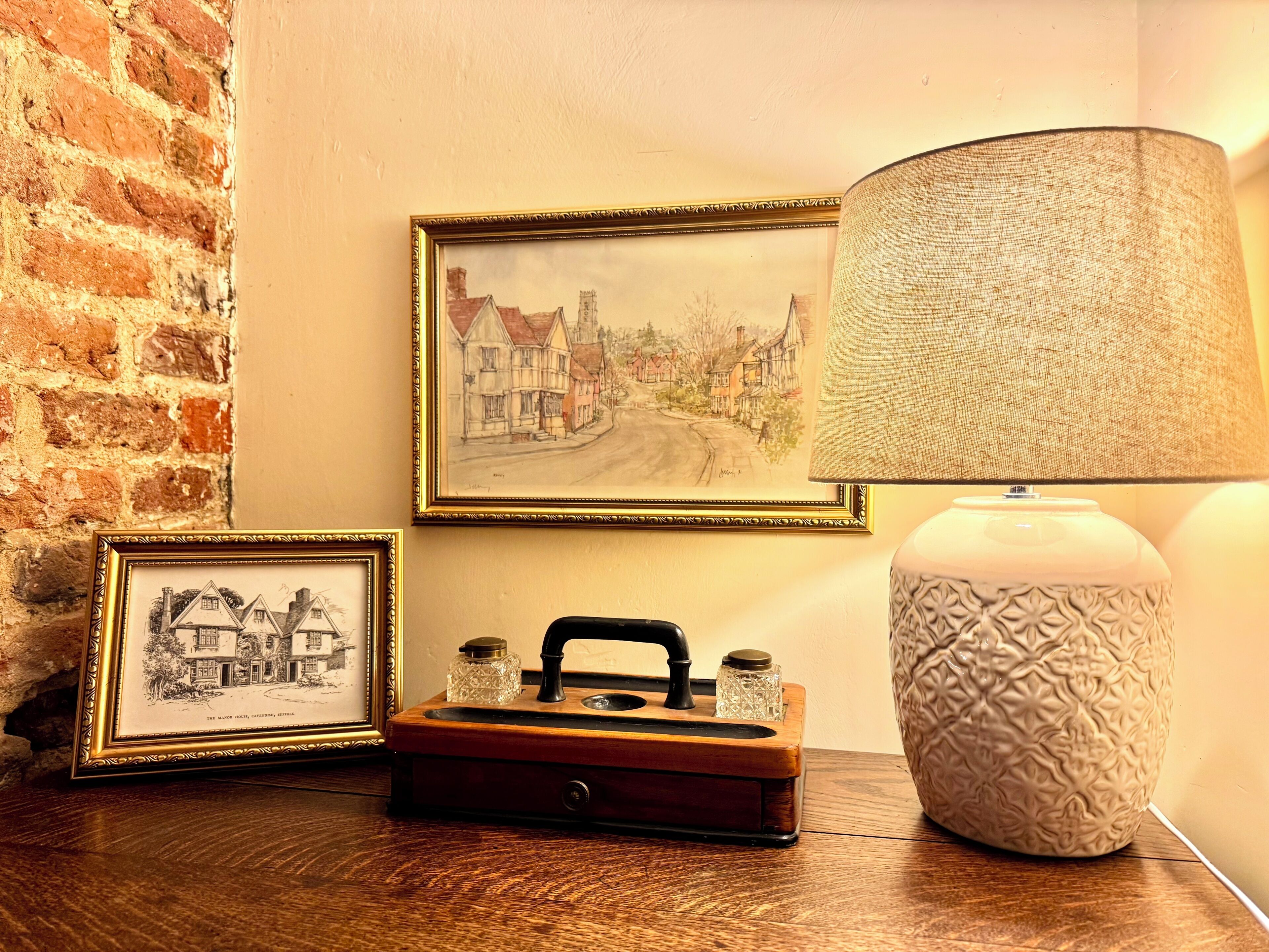 A framed painting, a vintage radio, and a lamp on a wooden table against a brick wall.
