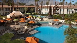 Ein Hotelpoolbereich mit mehreren Liegestühlen und Sonnenschirmen, umgeben von Palmen und Gebäuden.