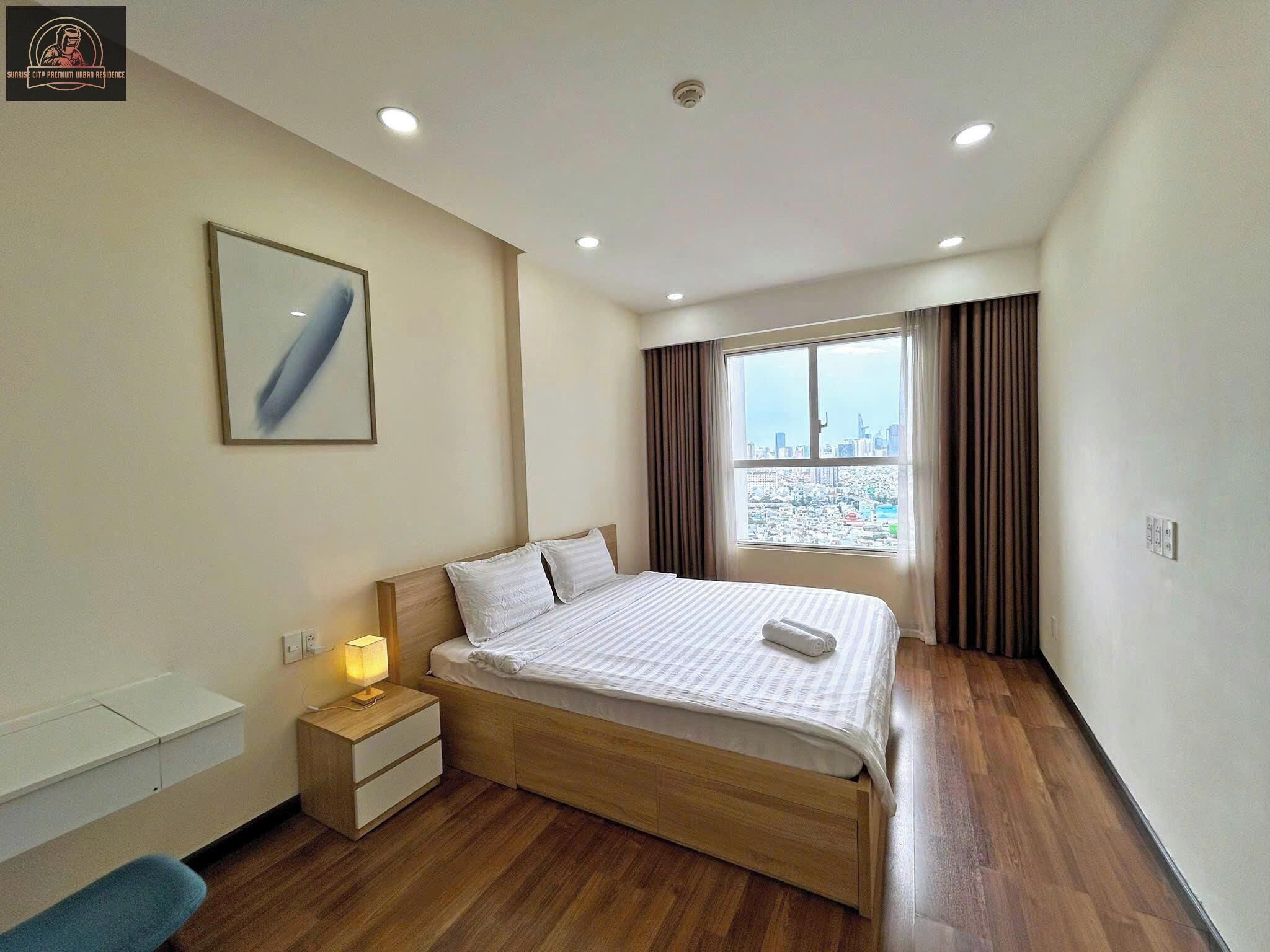 Habitación de hotel con cama, mesita de noche, lámpara y vista a la ciudad por la ventana.