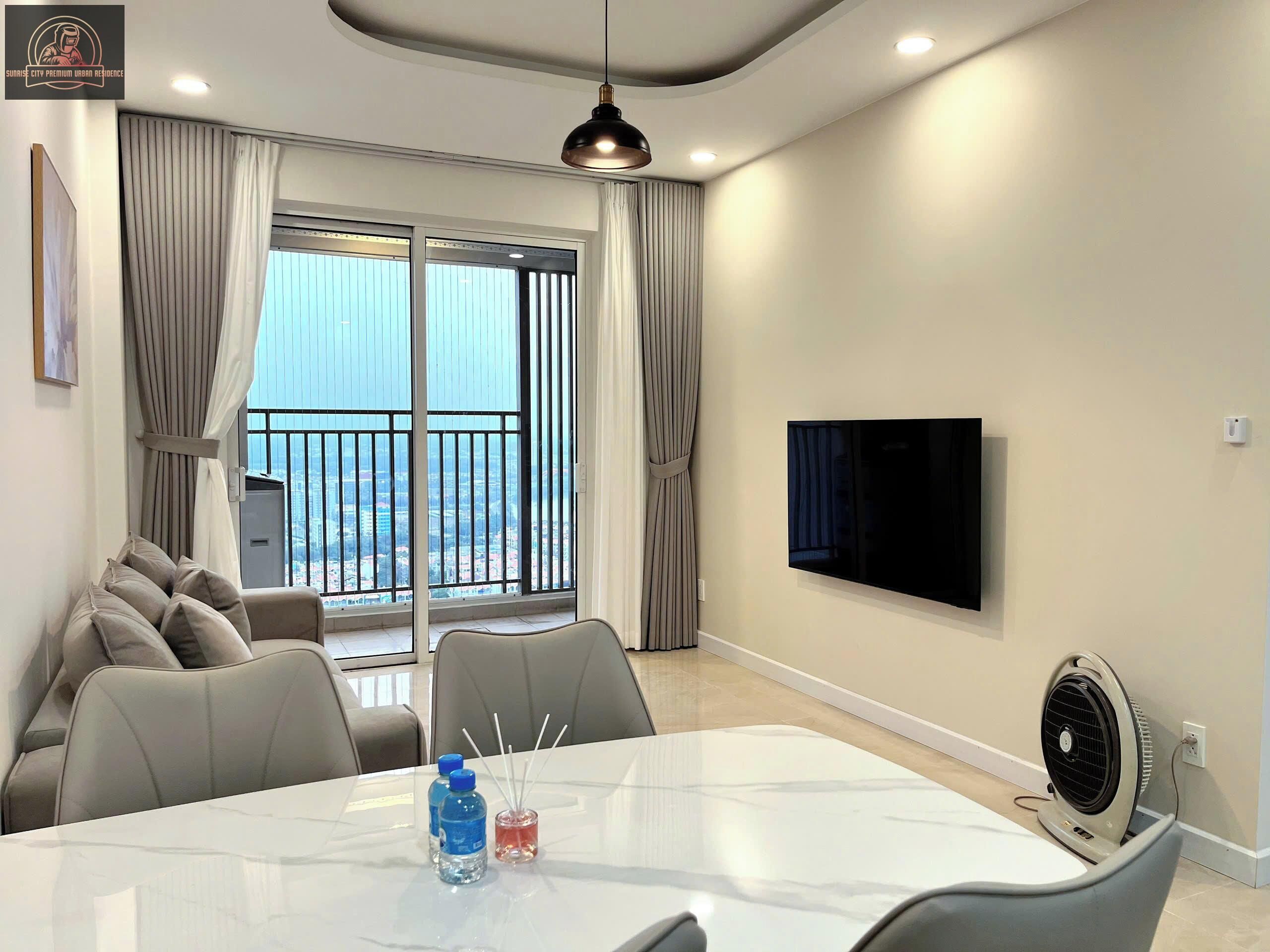 A modern living room with a large TV, comfortable seating, a dining table, and a balcony with a city view.