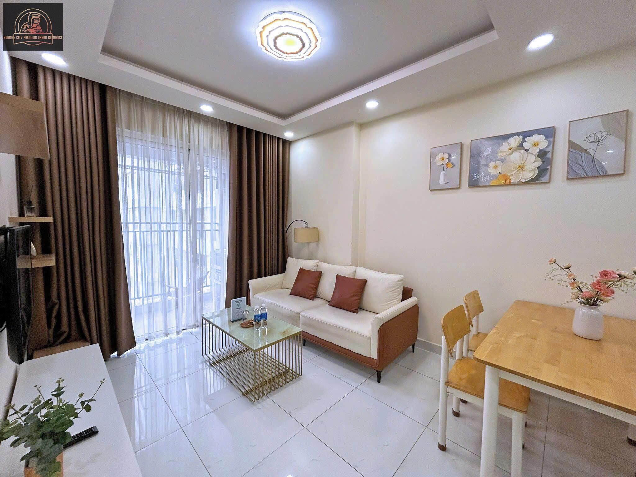 A modern living room with a white sofa, a glass coffee table, and a wooden dining table. There is a TV on the left, a lamp on the right, and a plant on the floor.