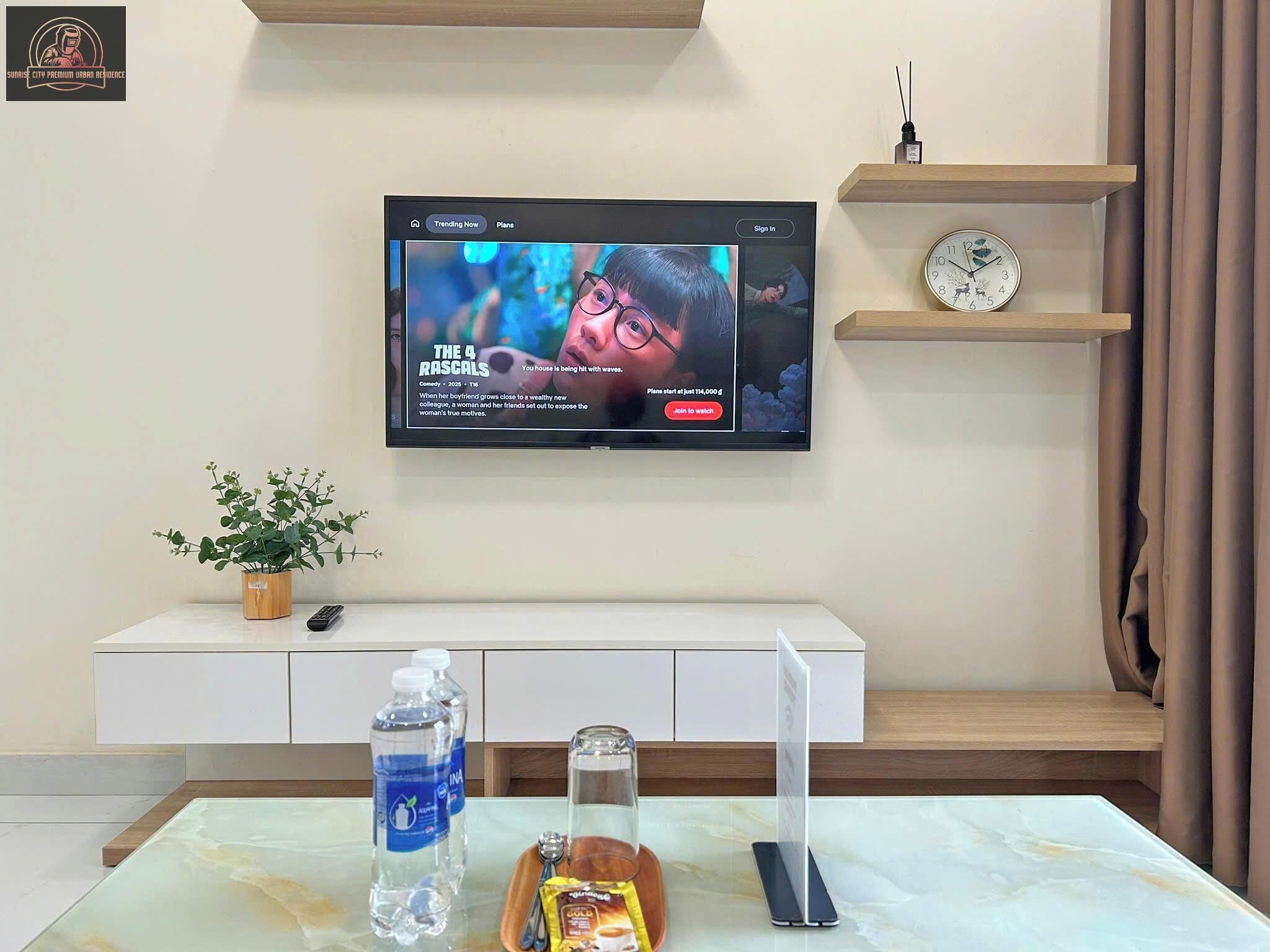 Una sala de estar moderna con un televisor de pantalla plana montado en la pared mostrando un cartel de película. Hay una mesita blanca con una planta en maceta, una botella de agua, una taza de café y un teléfono inteligente. Un reloj está sobre una estantería de madera encima de la mesita.