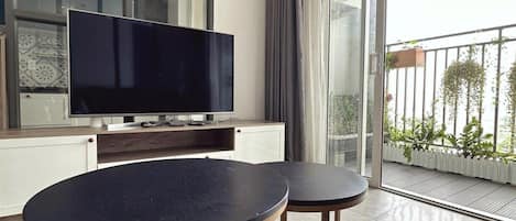 A modern living room with a large flat-screen TV, two round tables, and a balcony with plants.