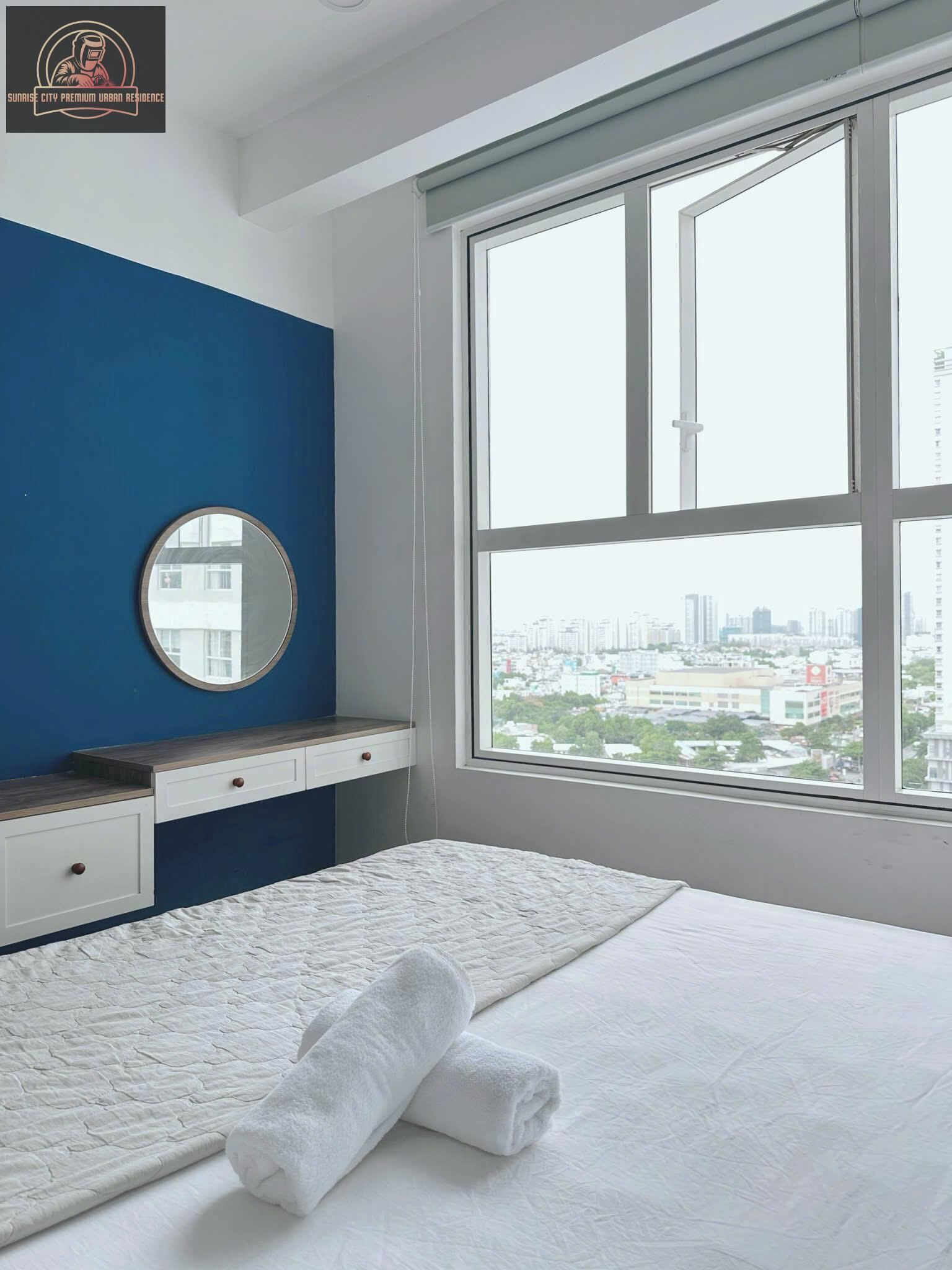 A bedroom with a large window, a round mirror, a bed with white bedding, and a blue accent wall.