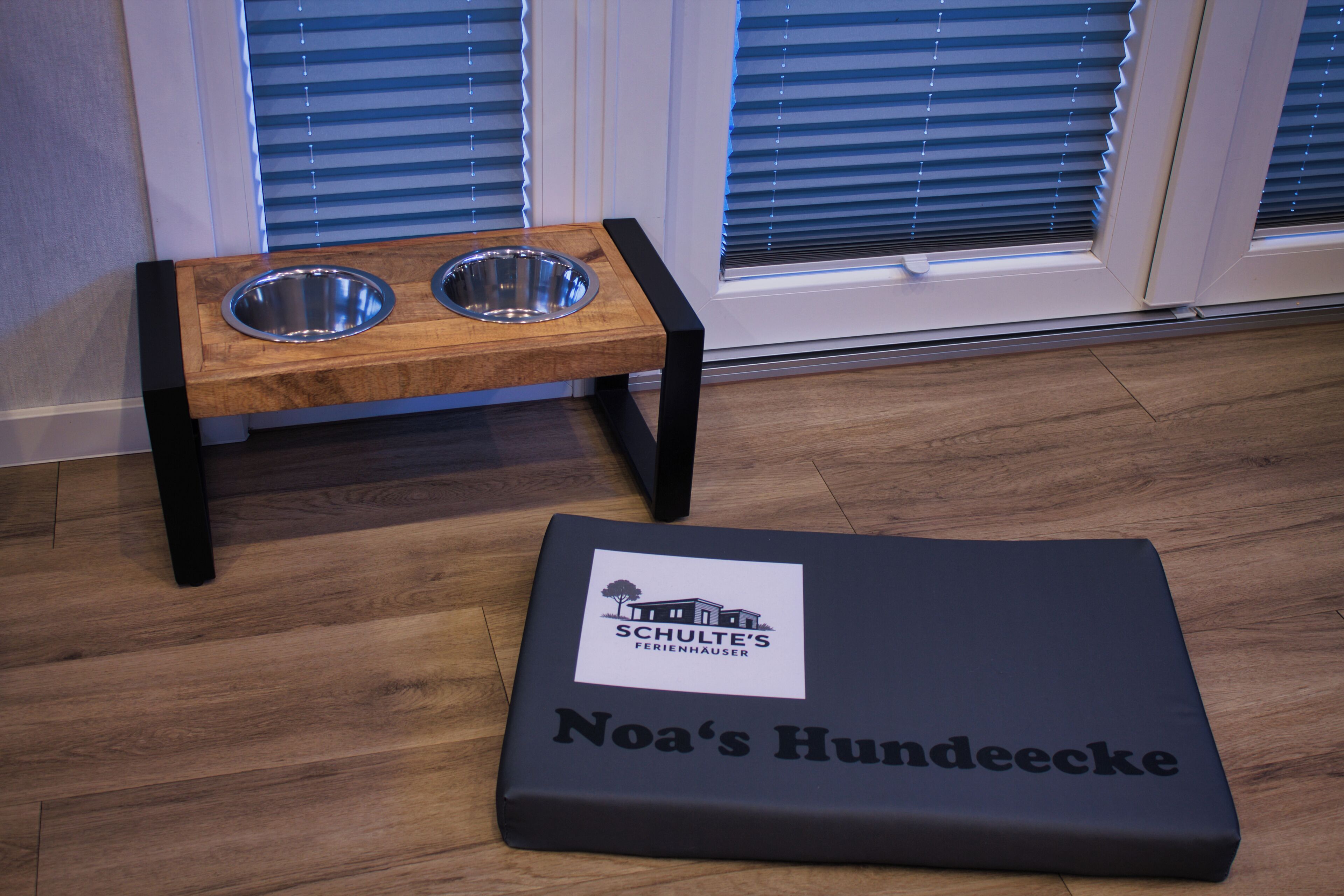 Ein Haustierbett mit dem Aufdruck "Noa's Hundeecce" und eine Holz-Futterstation mit zwei Metallnäpfen.