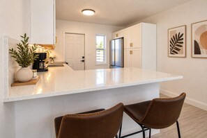 Une cuisine moderne avec un plan de travail blanc, deux chaises en cuir marron, un réfrigérateur et une machine à café.