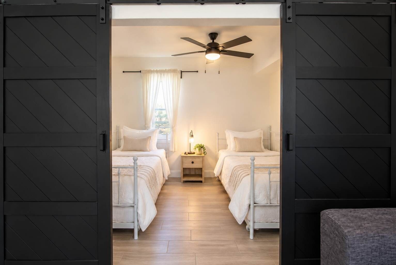 Une chambre avec deux lits, un ventilateur de plafond, une table de chevet avec une lampe et une fenêtre avec des rideaux.