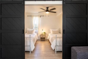 A bedroom with two beds, a ceiling fan, a bedside table with a lamp, and a window with curtains. - Sea Glass Beach Compound | 10BR, Sleeps 28, Near Beach | By Golden Coast Rentals (Clearwater)