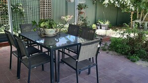 Mesa con cubierta de vidrio y cuatro sillas, rodeada de plantas en macetas y una cerca verde.