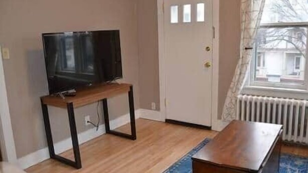 Een kamer met een houten vloer, een televisie op een standaard, een witte deur en een raam met uitzicht op gebouwen.