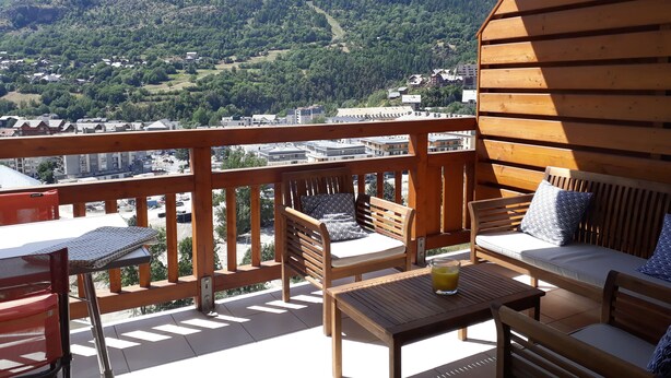 Eine Holzterrasse mit Gartenmöbeln, darunter eine Sitzecke und Stühle, mit Blick auf eine gebirgige Landschaft und eine darunterliegende Stadt.