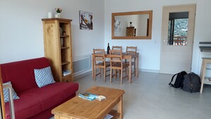 Ein Wohnzimmer mit rotem Sofa, Couchtisch aus Holz und Bücherregal. Es gibt einen Essbereich mit Holztischen und -stühlen, und einen Spiegel an der Wand.