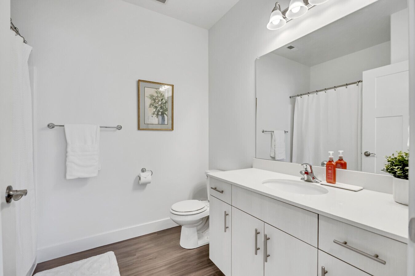 Un baño moderno con un lavamanos, espejo y retrete blancos. Hay un perchero, un cuadro en la pared y una barra para cortina de ducha.